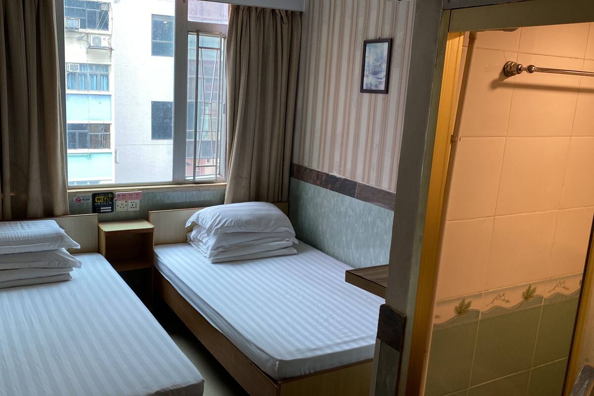 A hotel room with two single beds, a small wooden nightstand, a window with curtains, a wall-mounted picture, and a tiled shower area.