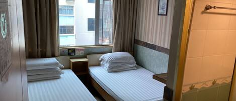 Habitación de hotel con dos camas individuales, una mesita de noche de madera, una ventana con cortinas, un cuadro colgado en la pared y una zona de ducha con azulejos.