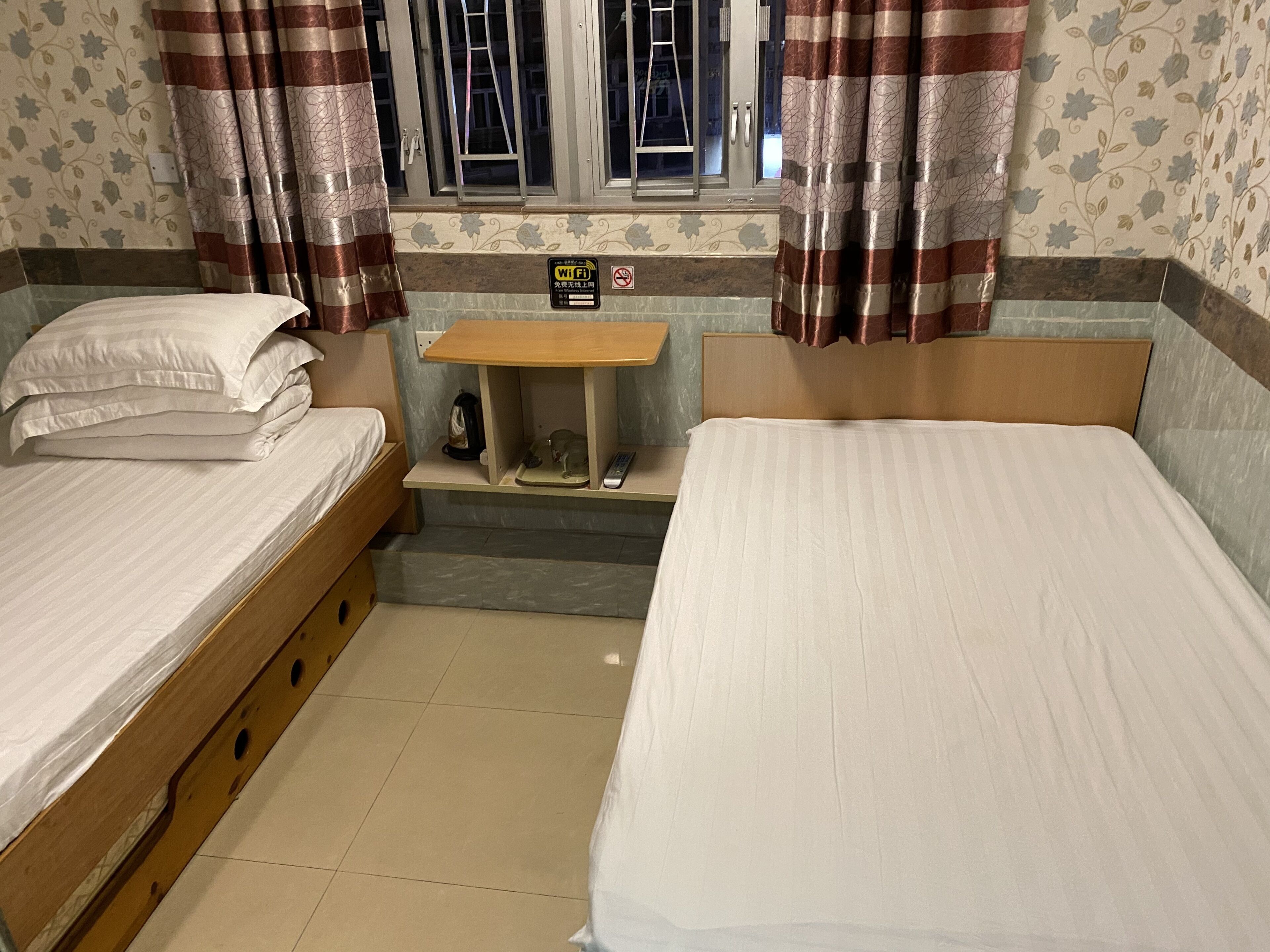 Ein Hotelzimmer mit zwei Einzelbetten, einem kleinen Holztisch, einem Fenster mit Vorhängen und einem Schild für WLAN.