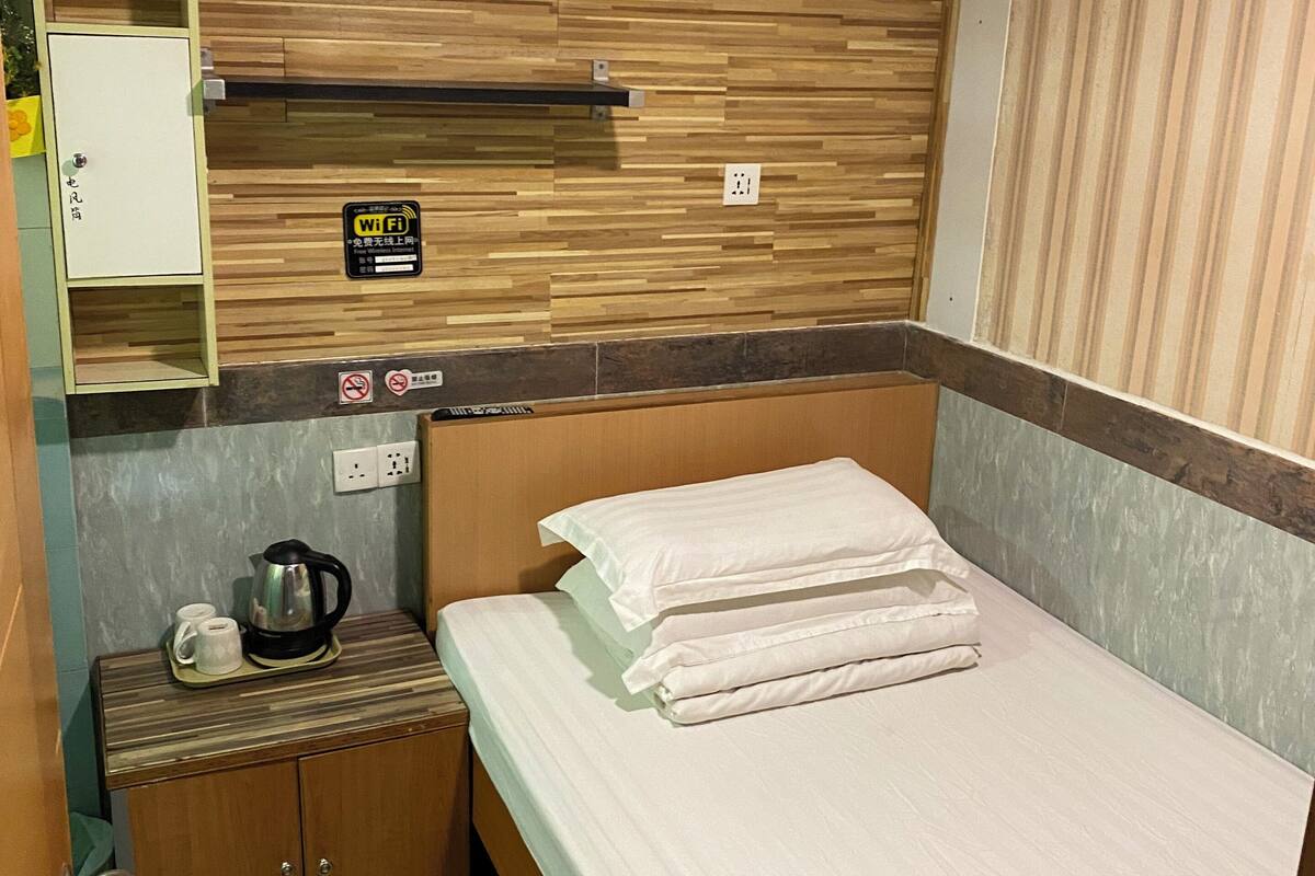 A hotel room with a wooden headboard, a bed with white linens, a small bedside table with a kettle and cups, a Wi-Fi sign, and a power outlet.