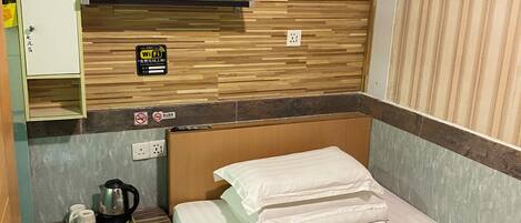 Habitación de hotel con cabecera de madera, cama con ropa de cama blanca, mesita de noche pequeña con tetera y tazas, señal de Wi-Fi y toma de corriente.