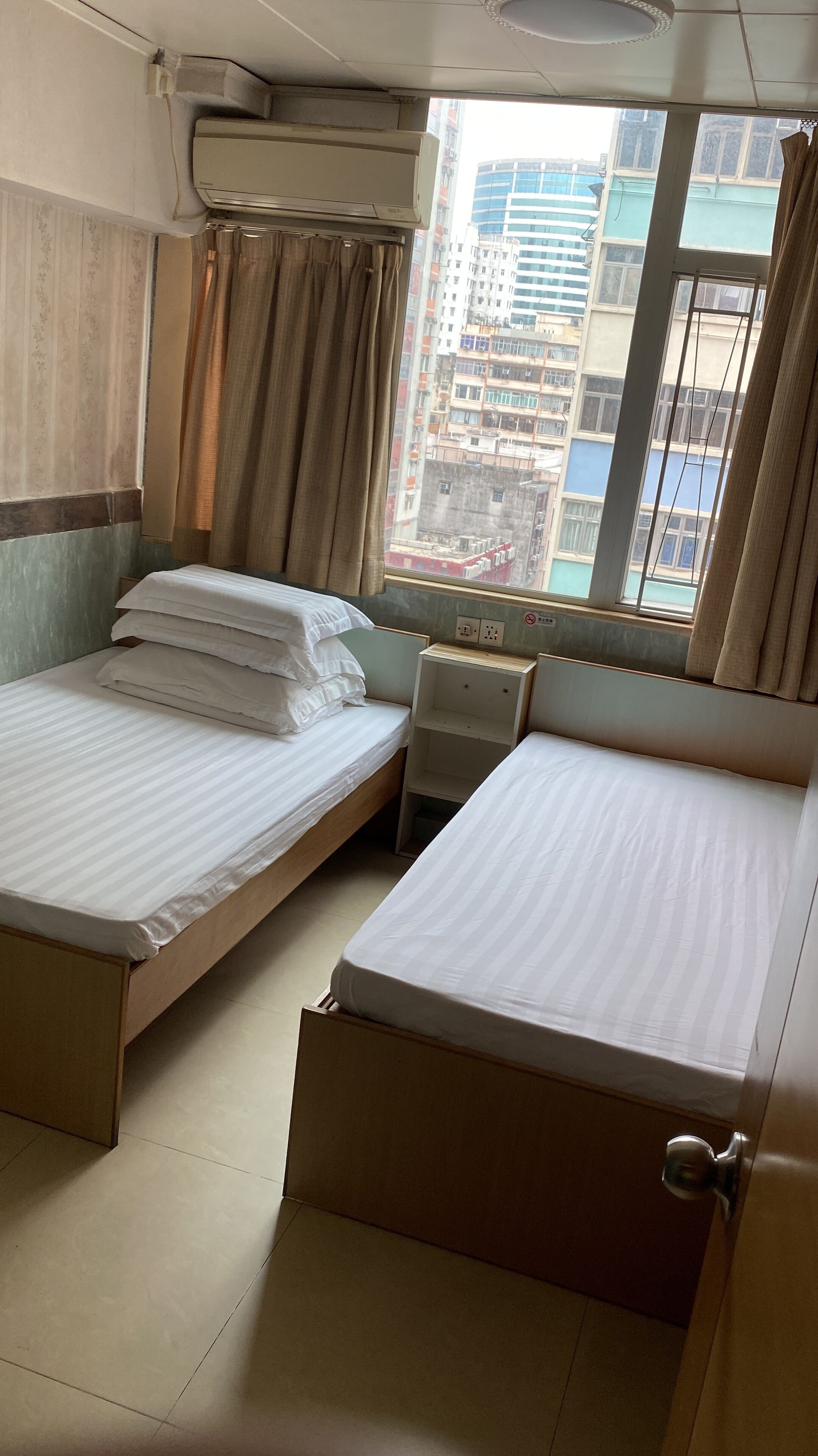 Een hotelkamer met twee bedden, een klein nachtkastje, een raam met gordijnen en uitzicht op gebouwen.