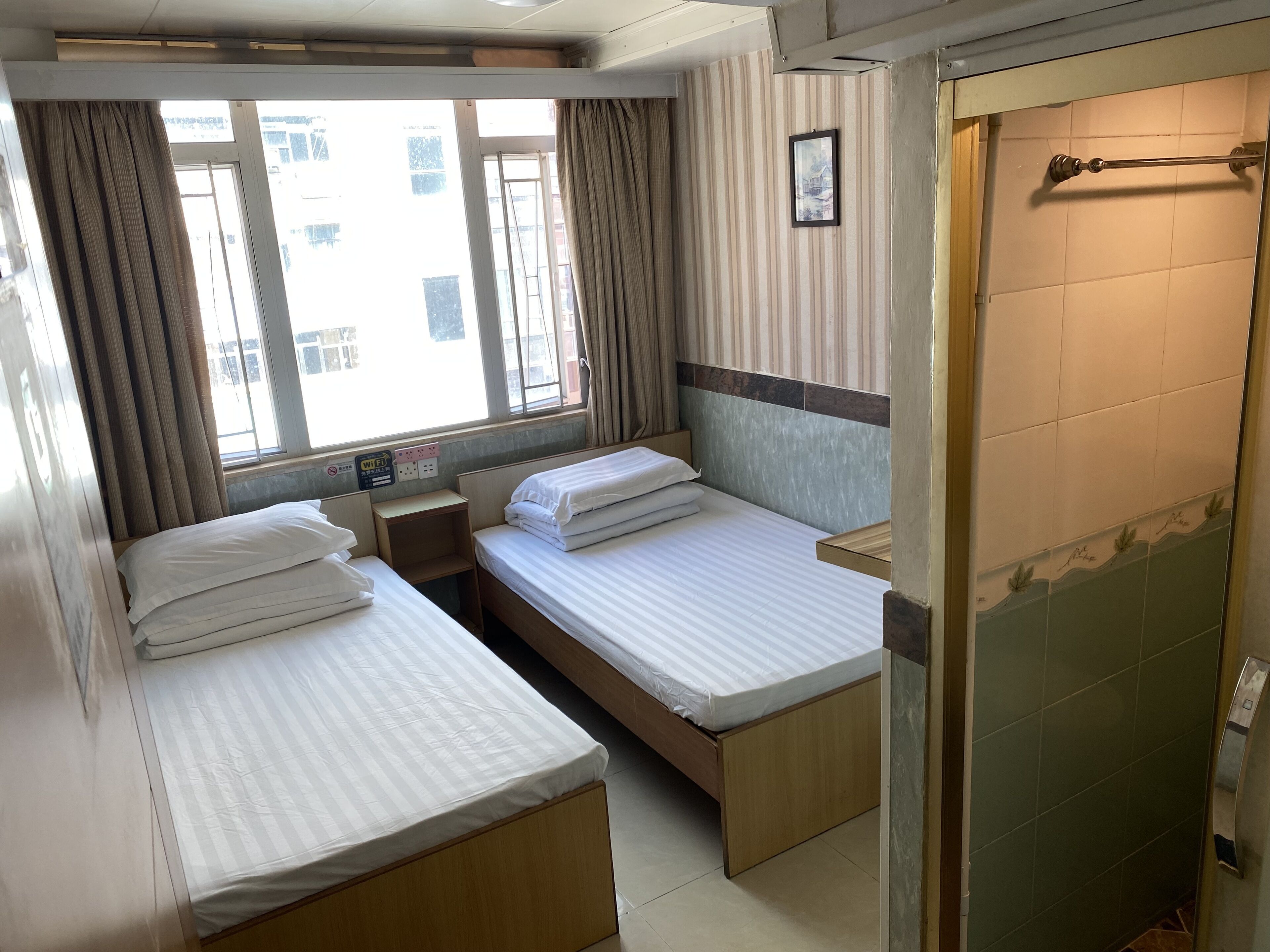A hotel room with two single beds, a small bedside table, a window with curtains, a shower area, and a wall-mounted picture.