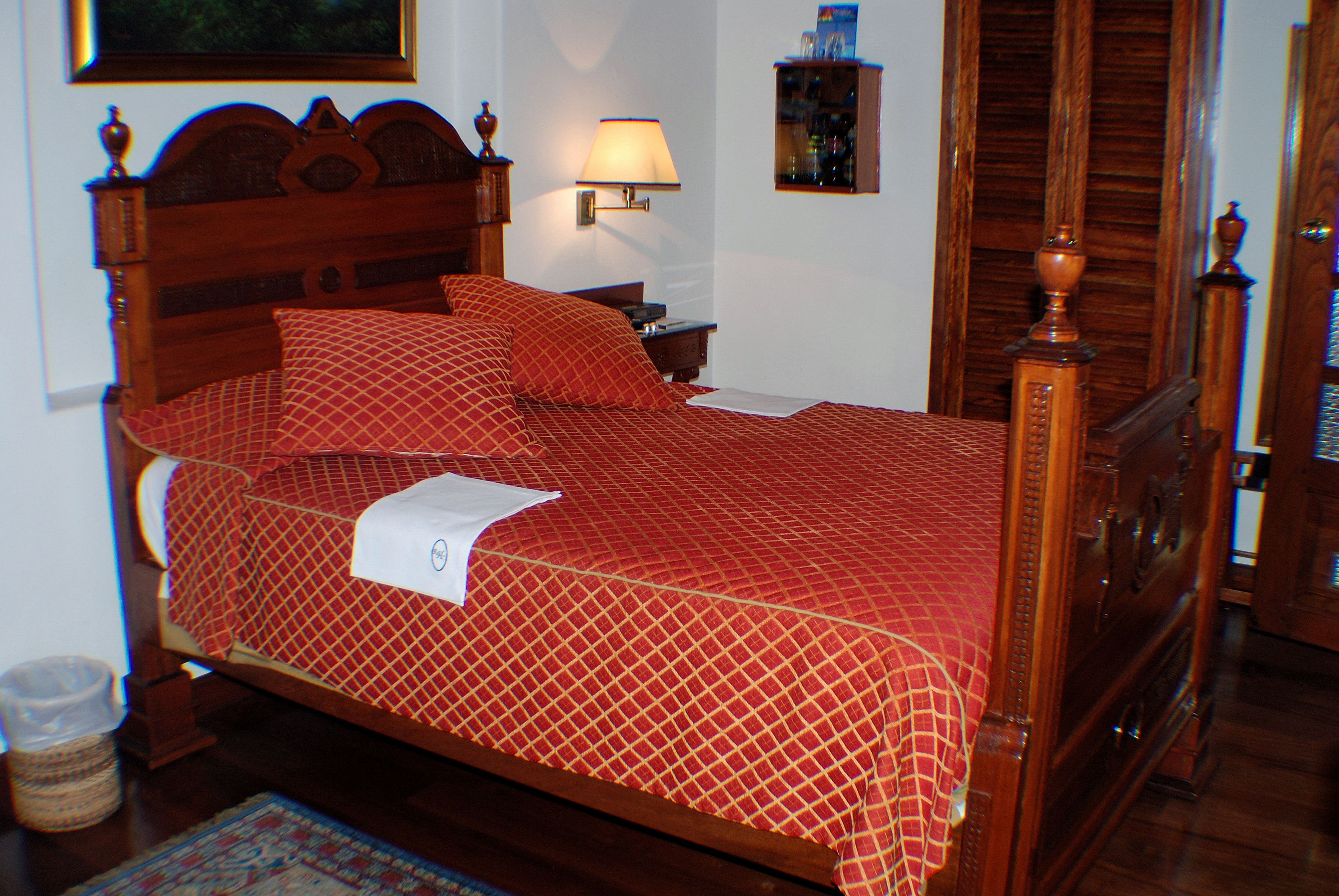 A neatly made bed with a red patterned bedspread and pillows, a bedside table with a lamp, and a wooden headboard.