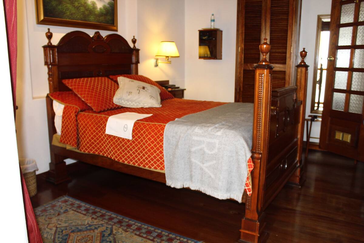 A traditional wooden bed with a red and white patterned bedspread, a white pillow, and a white blanket. There is a wooden nightstand with a lamp, a small mirror, and a bottle of water on the wall. The room has wooden flooring and a patterned rug.