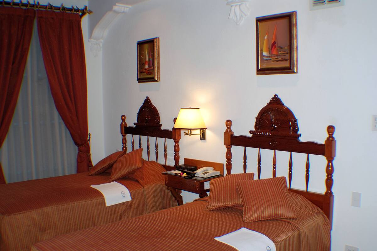 A hotel room with two beds, a nightstand, a lamp, and two paintings on the wall.
