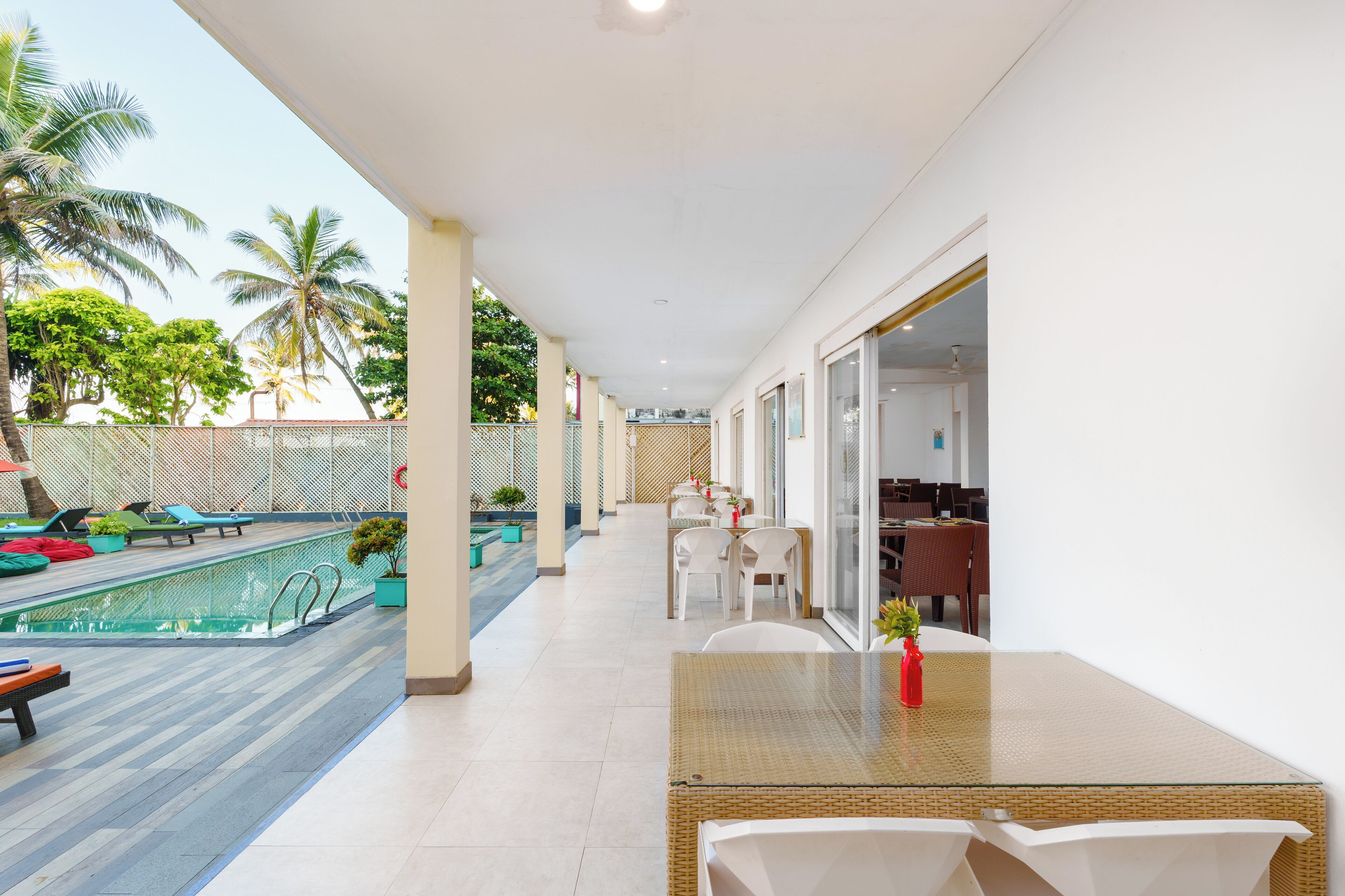 A poolside area with a table and chairs, a pool, and a covered seating area with a view of palm trees.