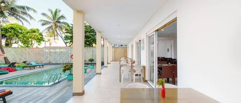 A poolside area with a table and chairs, a pool, and a covered seating area with a view of palm trees.