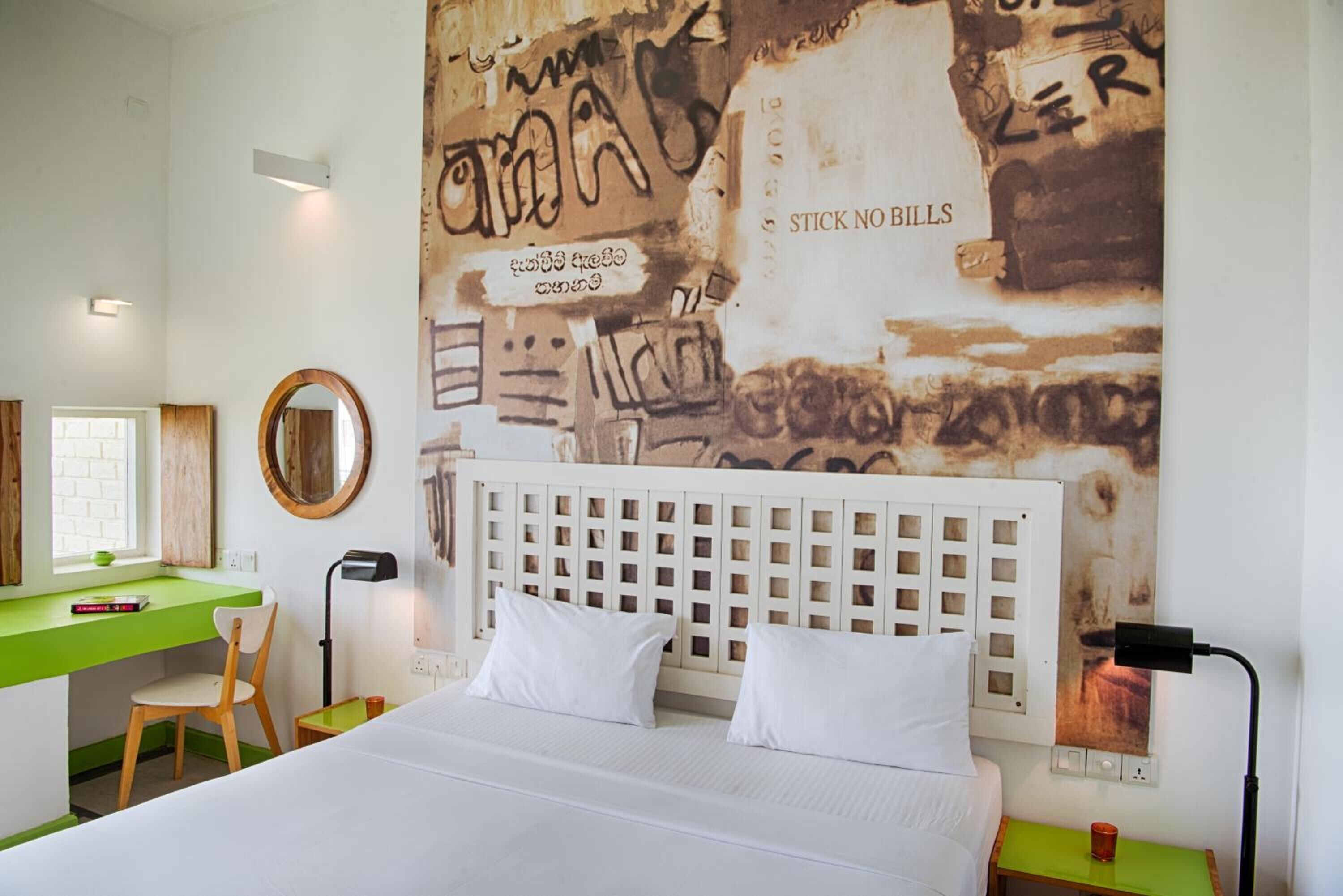 A bedroom with a bed, a desk, and a chair. There is a wall with graffiti and a sign that says "STICK NO BILLS".
