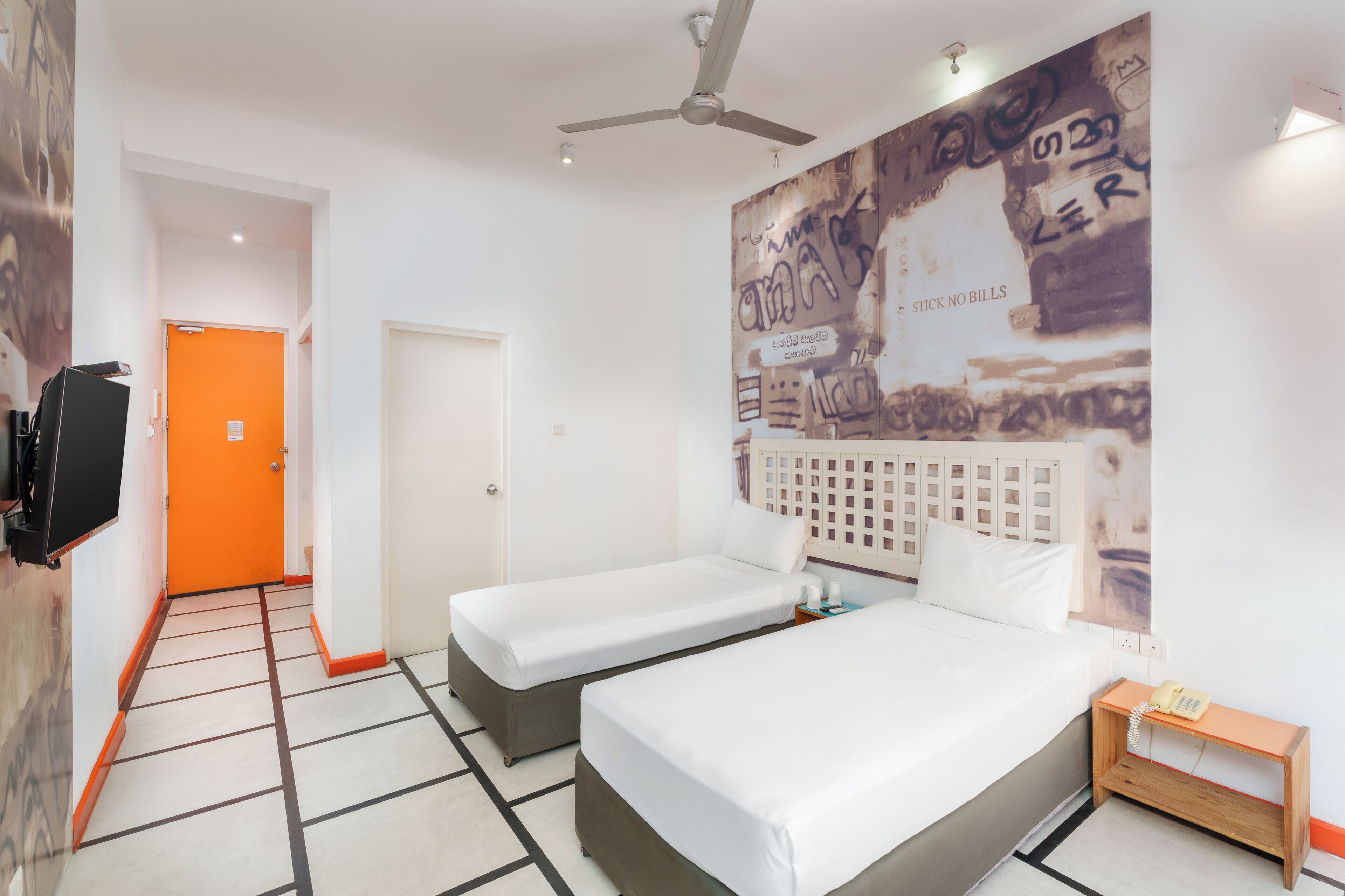 A hotel room with two single beds, a ceiling fan, a wall-mounted TV, and a mural with graffiti.