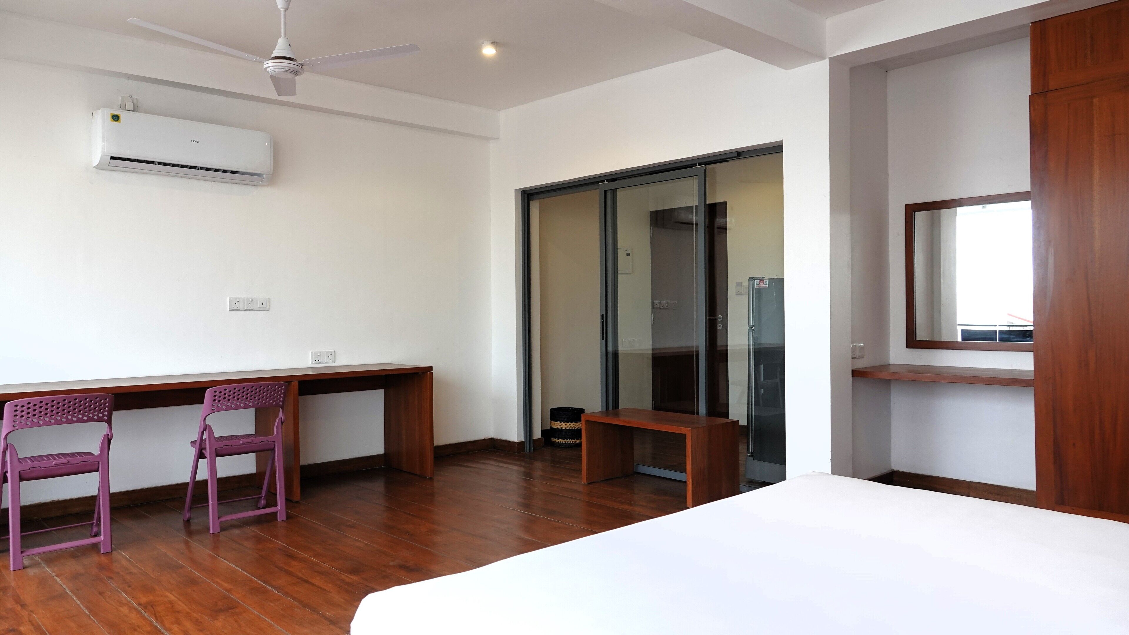A modern hotel room with a wooden desk, two purple chairs, a bed, a ceiling fan, an air conditioner, a sliding glass door, and a window with a view.