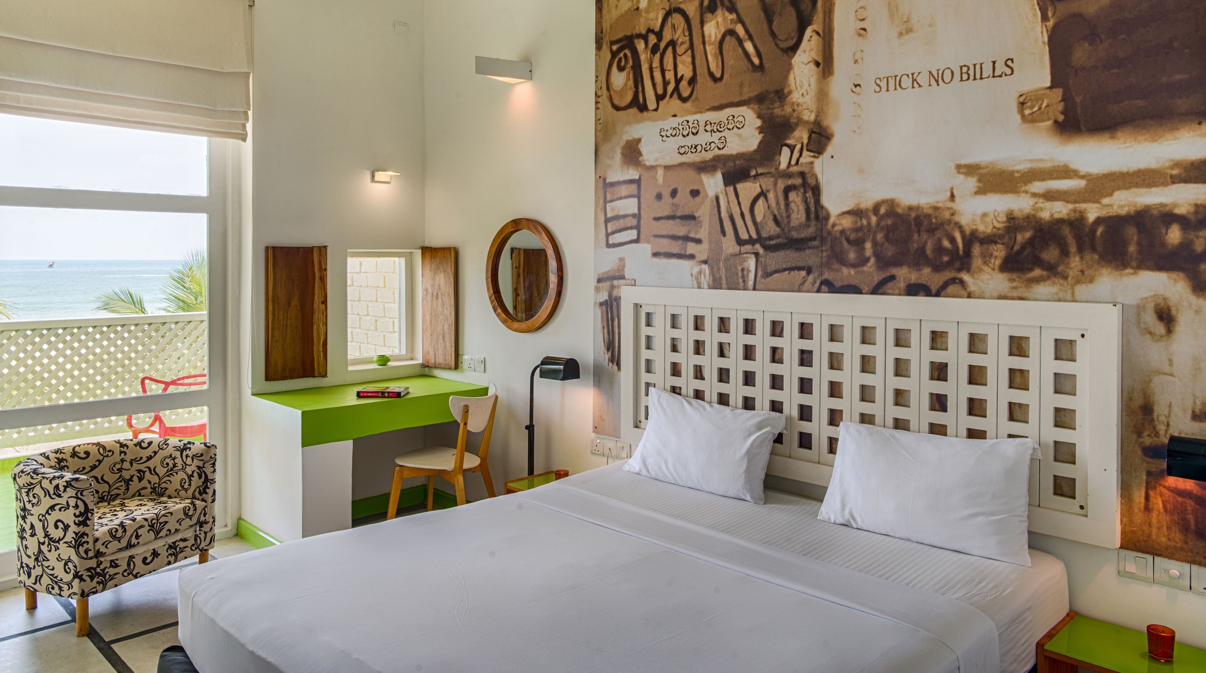 A hotel room with a bed, a desk, a chair, a window with a view of the sea, and a wall with graffiti and a sign that says "STICK NO BILLS".
