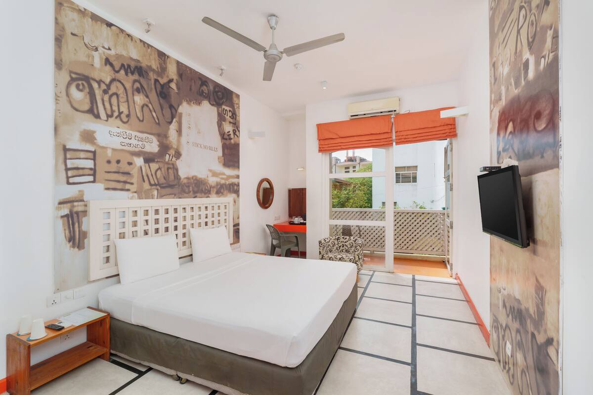 A bedroom with a large bed, a ceiling fan, a desk with a chair, and a balcony with a view.