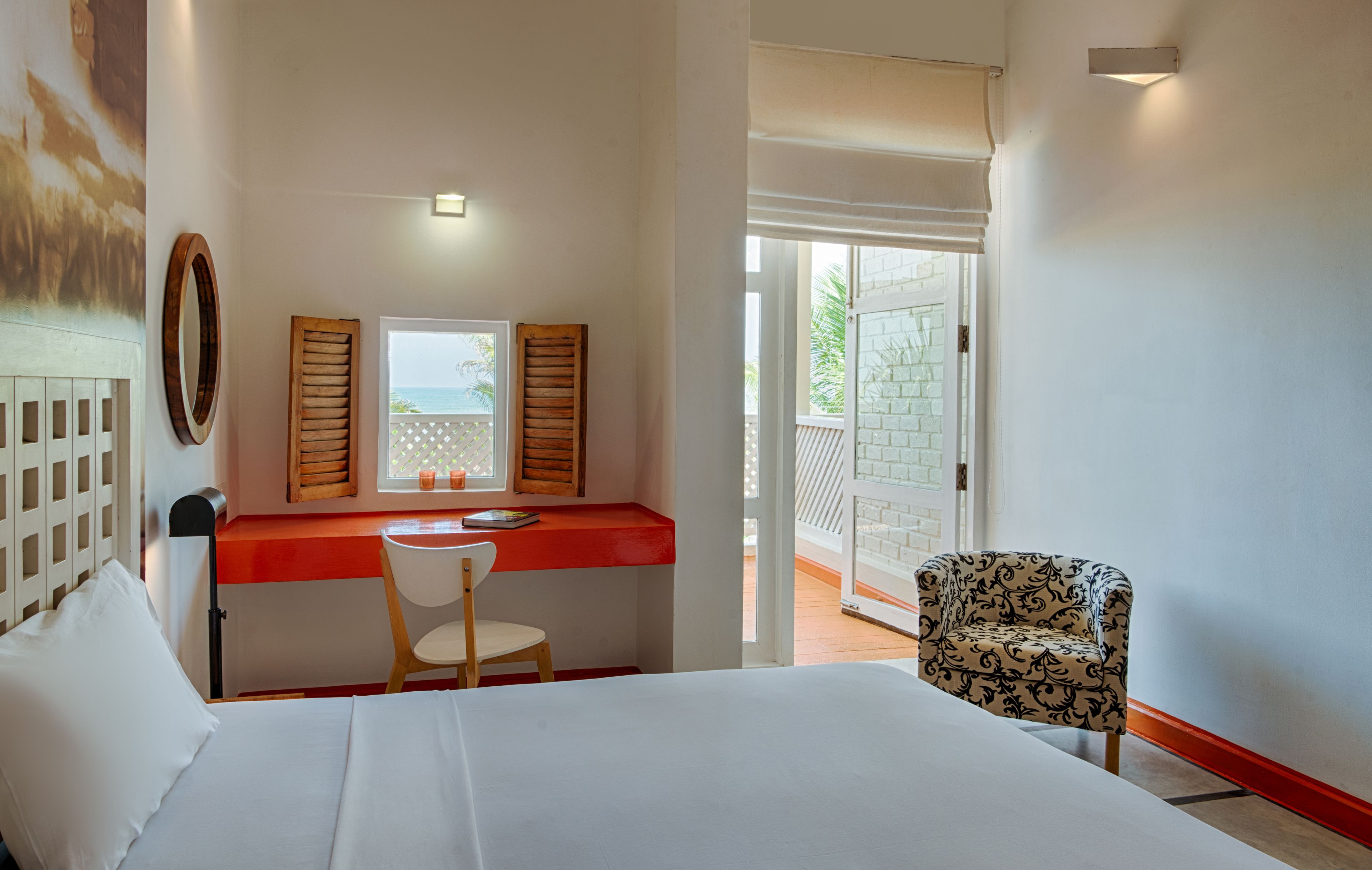 A bedroom with a bed, a desk with a chair, a window with shutters, and a view of the outside.