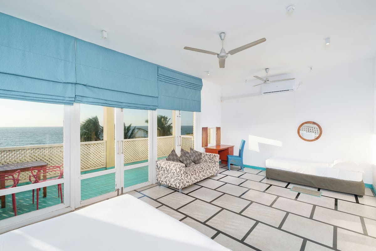 A room with a large window overlooking the sea, a ceiling fan, and a bed.
