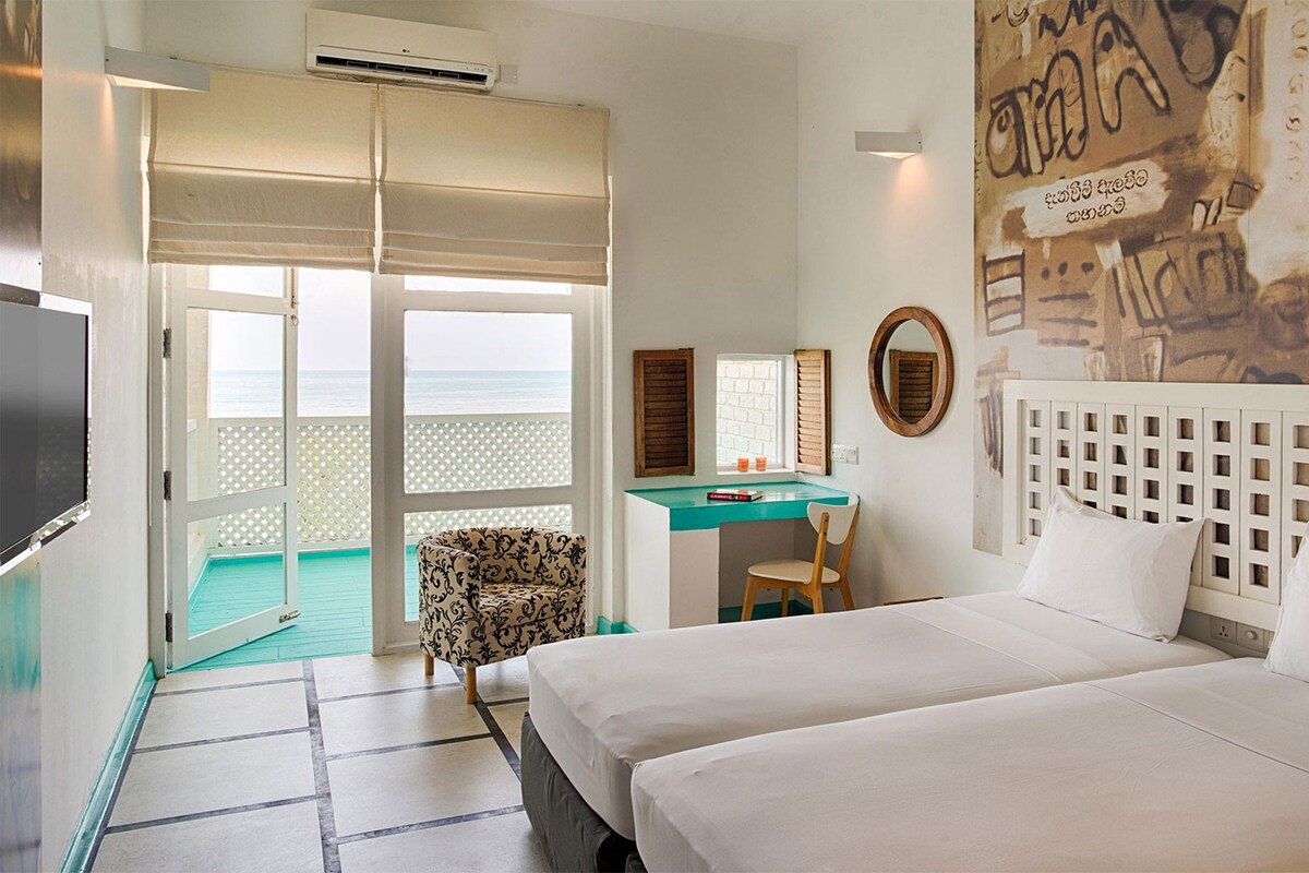 A hotel room with a bed, a desk, a chair, a TV, and a balcony with a view of the sea.
