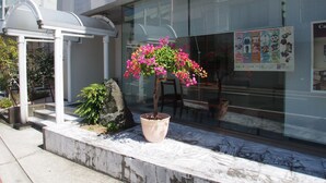 A building with a glass facade, a covered outdoor seating area, and a potted flowering plant. - Hotel Redent Ito (Ito)