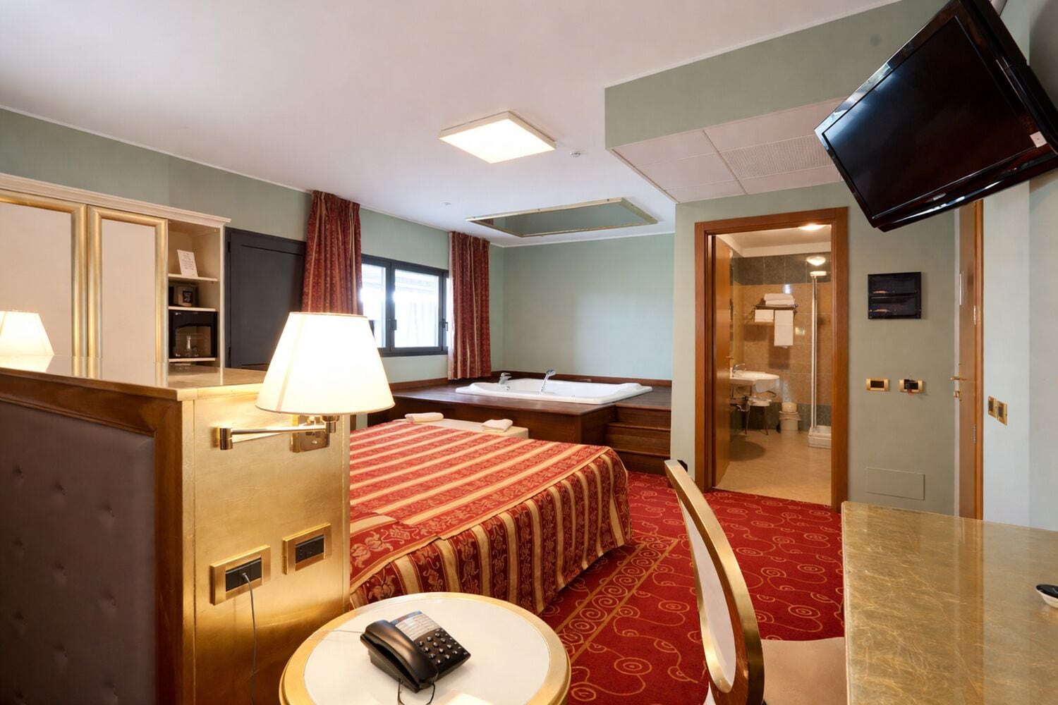 Habitación de hotel con cama, escritorio con lámpara, teléfono, televisión montada en la pared y baño visible a través de una puerta abierta.