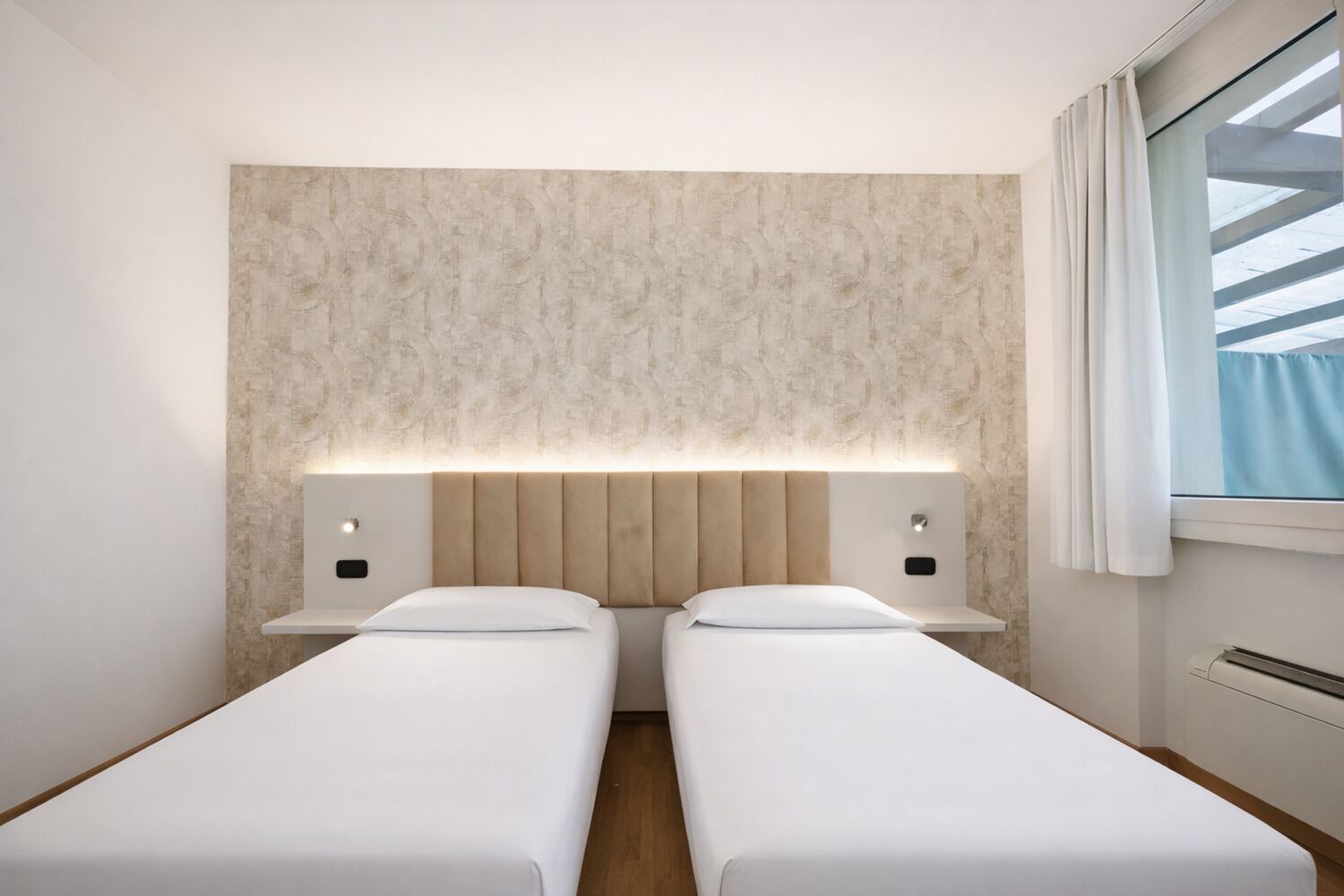 A hotel room with two single beds, a headboard with integrated lighting, a window with curtains, and a wall-mounted air conditioning unit.