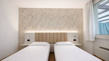 A hotel room with two single beds, a headboard with integrated lighting, a window with curtains, and a wall-mounted air conditioning unit.