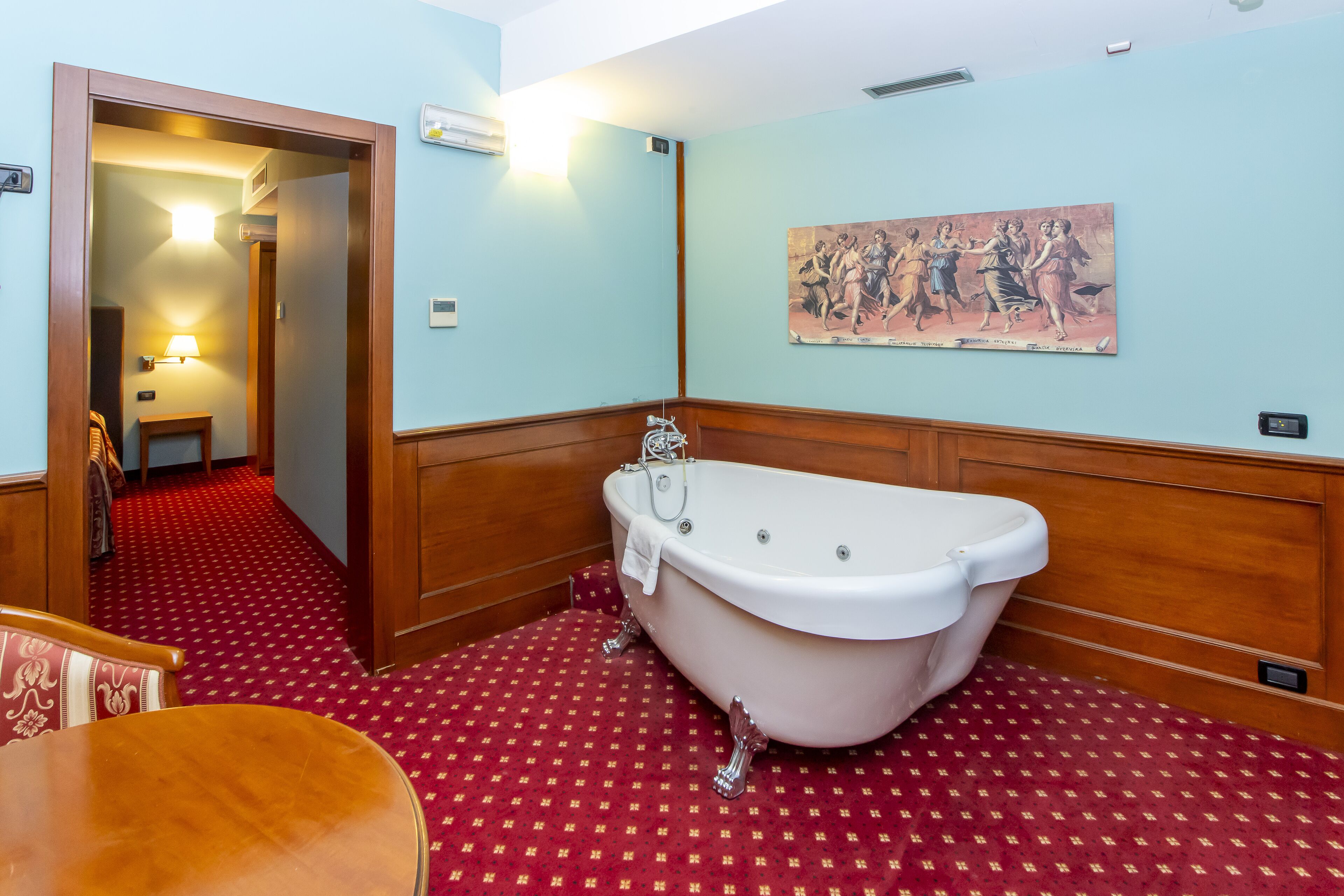 A bathroom with a bathtub, wooden paneling, a red patterned floor, a painting on the wall, and a door leading to another room.