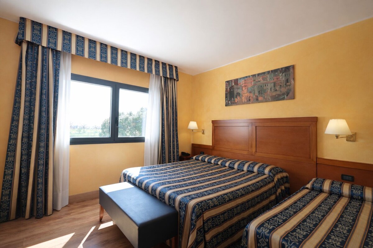 Ett hotellrum med en stor säng, en bänk, två sängbord, en tavla på väggen och gardiner.