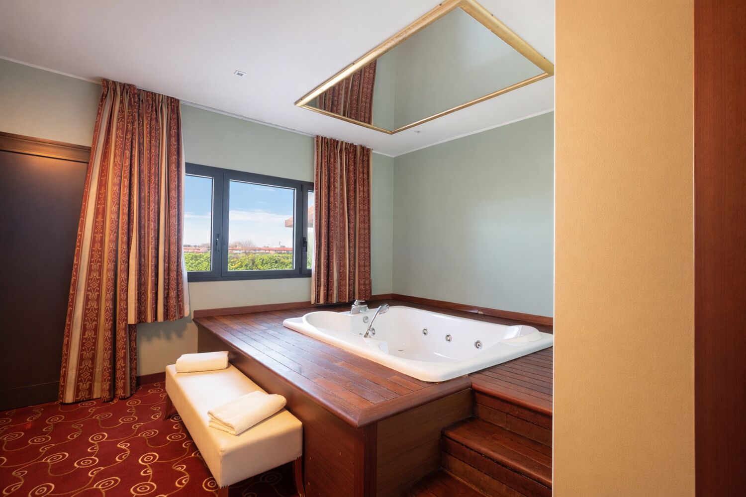 A bathroom with a large bathtub, a bench, and a window with curtains.
