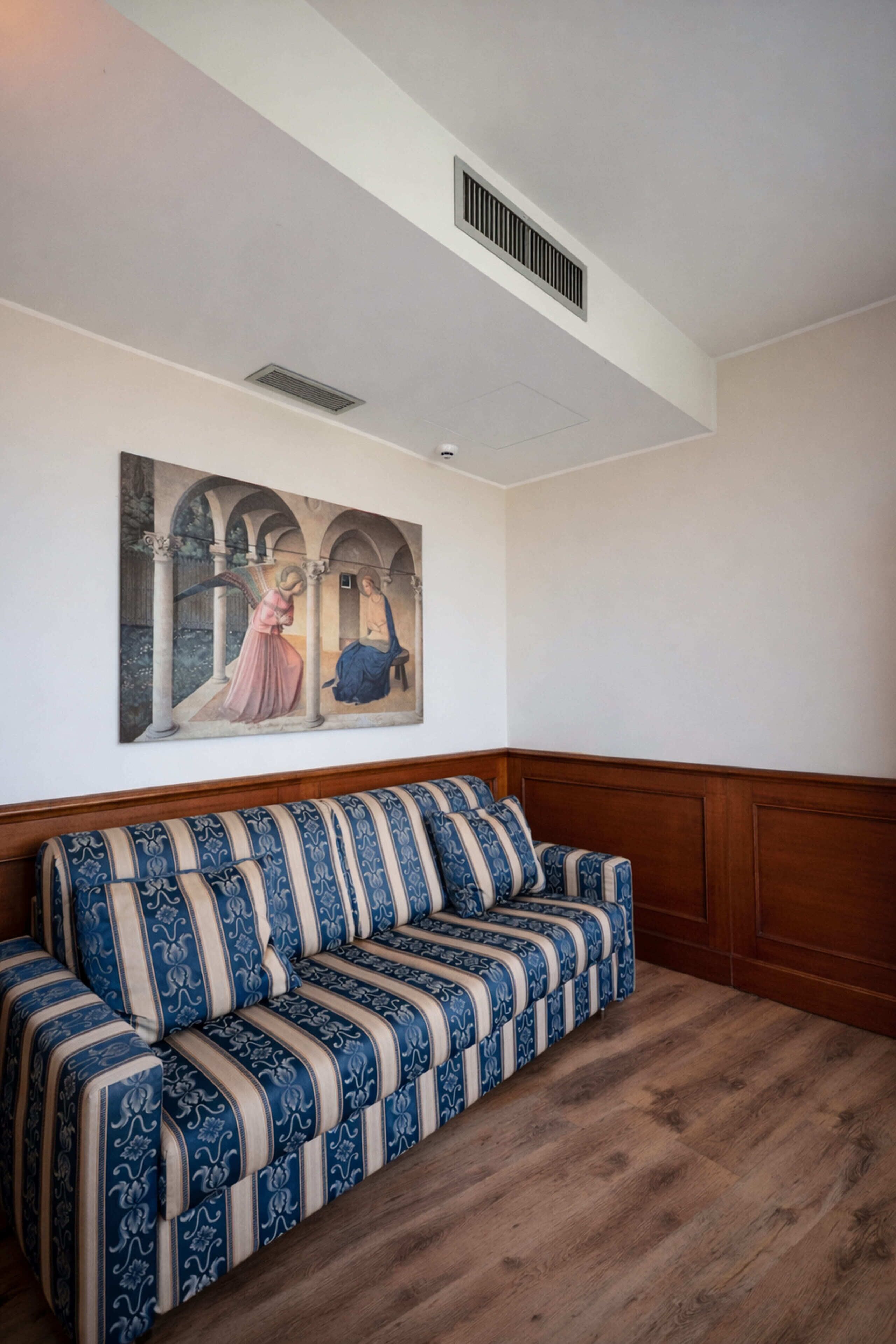A room with a wooden bench, a ceiling vent, and a wall-mounted painting.
