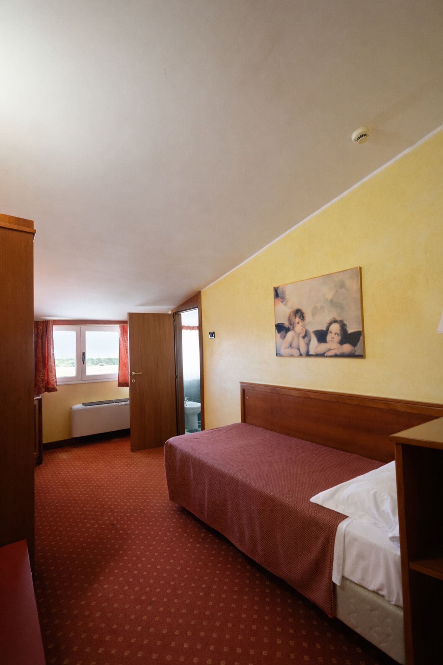 A hotel room with a bed, a window, a bathroom, and a painting on the wall.