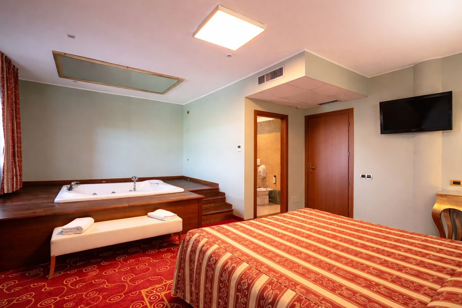 Habitación de hotel con una cama grande, una zona de estar con un banco, una bañera y un televisor en la pared.