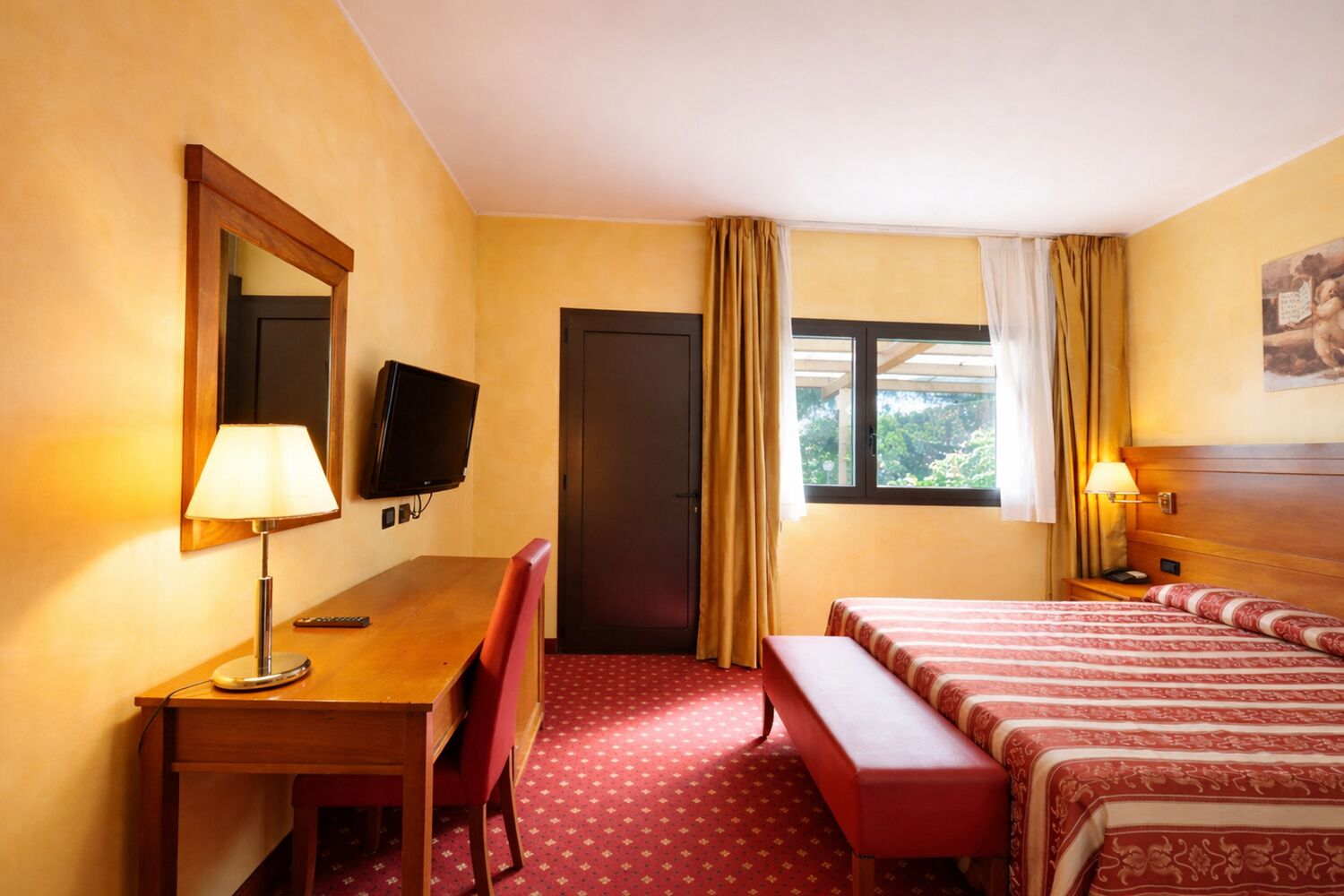A hotel room with a desk, a television, a bed with a patterned bedspread, a bench, and a window with curtains.