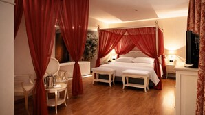 A spacious bedroom with a large bed, a sitting area with a table and chairs, and a television. - LVG Hotel Collection – Riz (San Genesio ed Uniti)