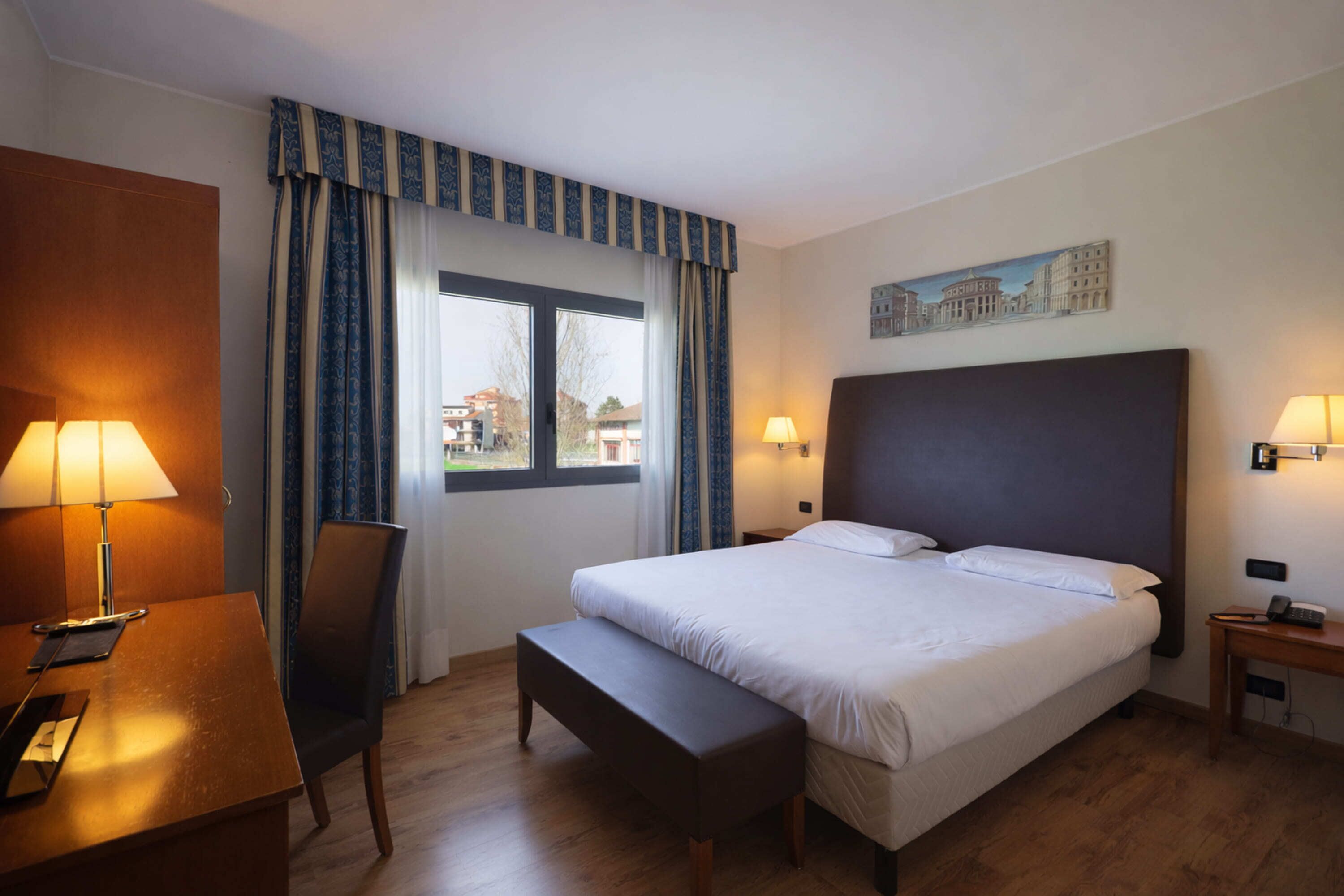 Une chambre d’hôtel avec un lit, un bureau avec une lampe, une chaise, un banc et une fenêtre avec des rideaux.