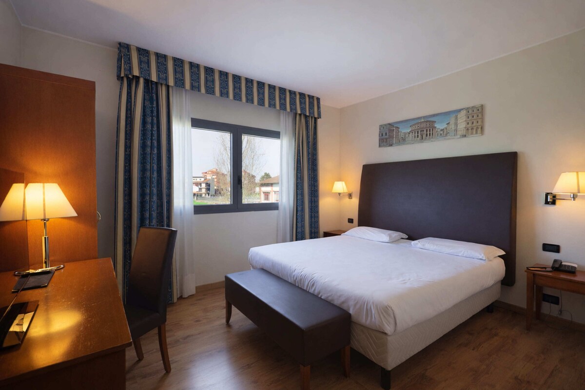 Ett hotellrum med en säng, ett skrivbord med en lampa, en stol, en bänk och ett fönster med gardiner.