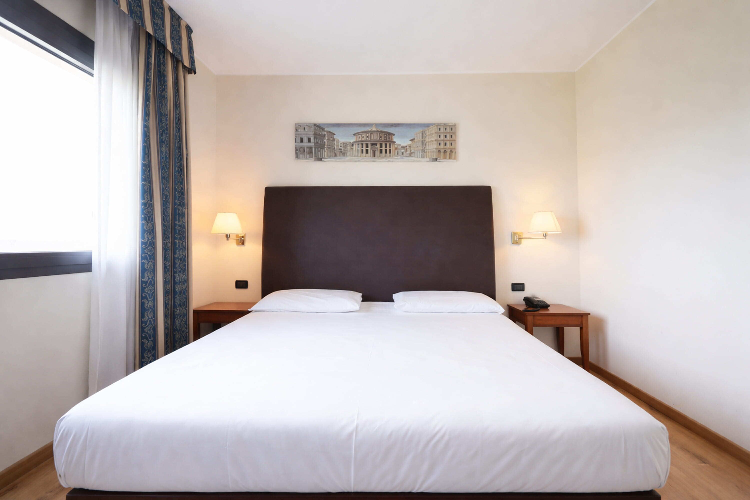 Een hotelkamer met een groot bed, twee nachtkastjes, een schilderij aan de muur en een raam met gordijnen.