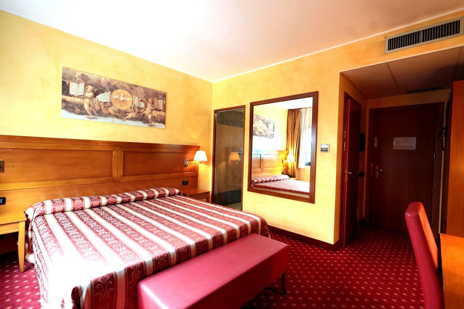 Une chambre d’hôtel avec une tête de lit en bois, un grand lit recouvert d’une couvre-lit à motifs, un banc, une table de chevet, un miroir et une porte.
