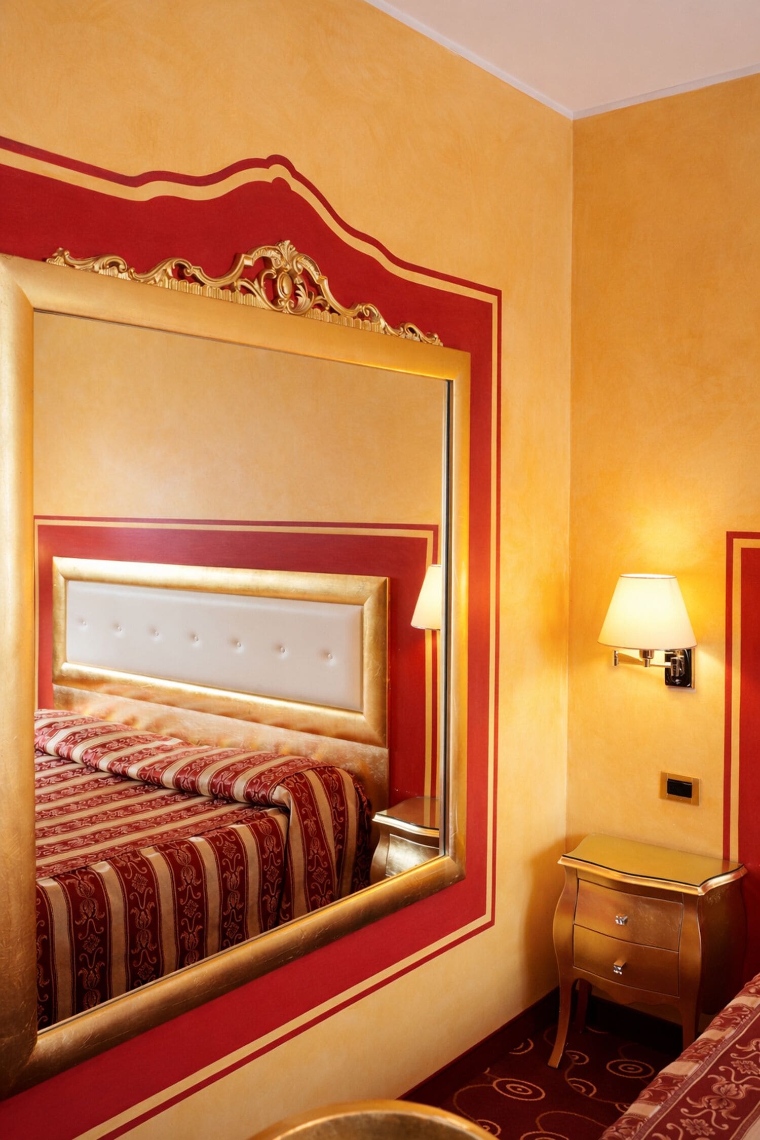 A hotel room with a large mirror, a bed with a red and gold headboard, a bedside table with a lamp, and a small nightstand.