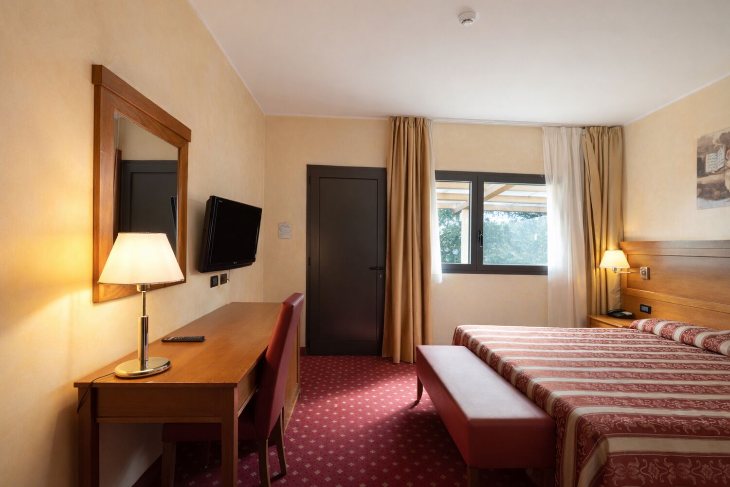 A hotel room with a desk, a television, a bed, a bench, and a window with curtains.