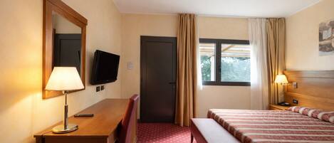A hotel room with a desk, a television, a bed, a bench, and a window with curtains.