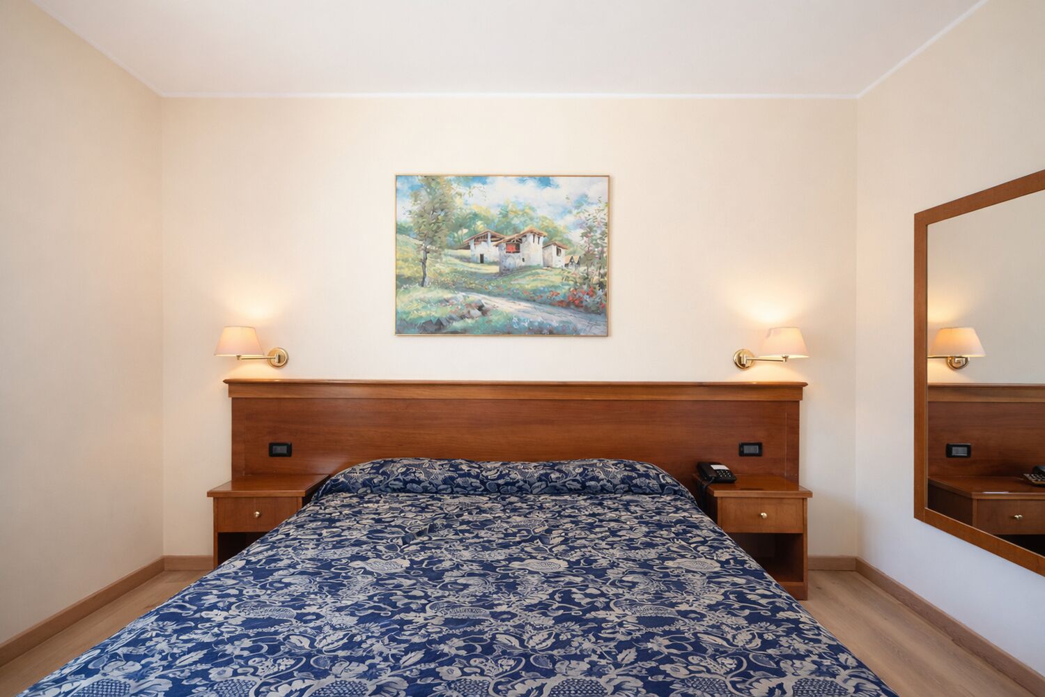 Ein Schlafzimmer mit einem hölzernen Kopfstück, einem Bett mit einem gemusterten Bettbezug, Nachttischlampen, einem Nachttisch und einem Bild an der Wand.