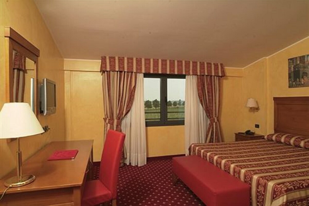 A hotel room with a bed, a desk, a chair, a lamp, and a window with curtains.