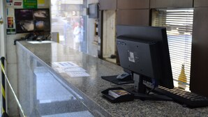 A reception desk with a computer monitor, keyboard, and mouse. There are signs on the wall and a window with blinds. - Hotel Scala (Porto Alegre)