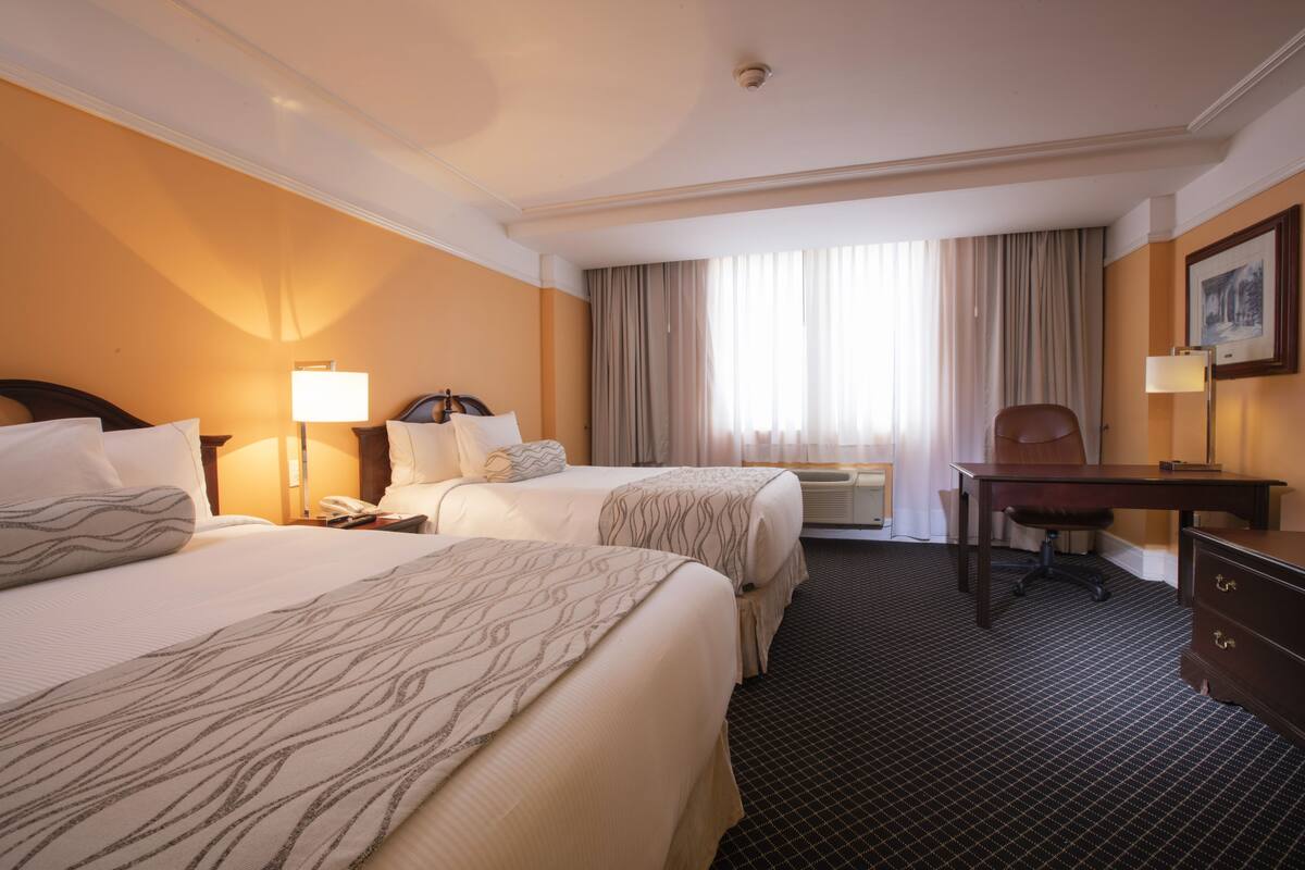 Ein Hotelzimmer mit zwei Betten, einem Schreibtisch, einem Stuhl, einem Telefon und einem Fenster mit Vorhängen.