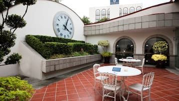 A courtyard with a clock, white chairs, and tables, surrounded by greenery and a building in the background.