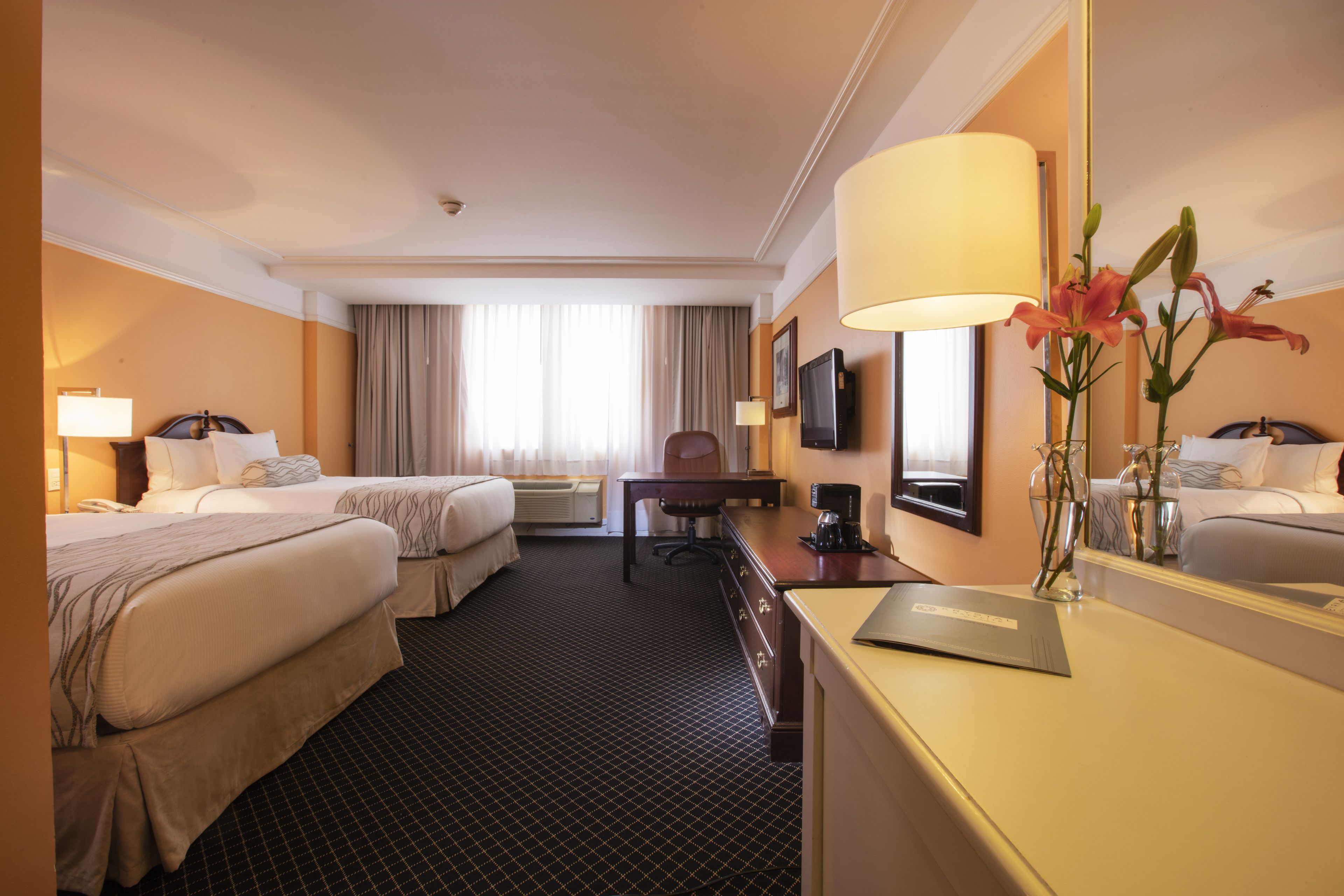 A hotel room with two beds, a desk, a chair, a television, and a large mirror.