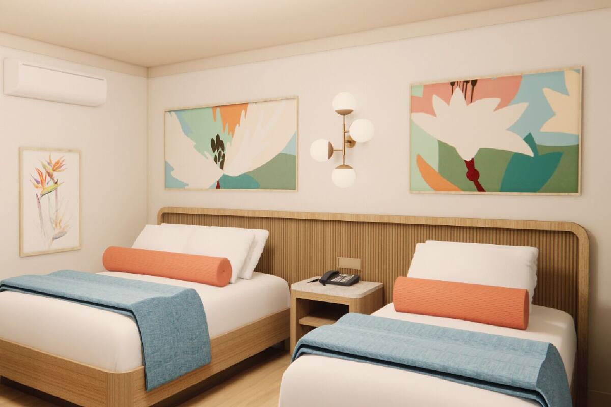 A hotel room with two beds, each with a wooden headboard, white bedding, and orange pillows. There are wall-mounted light fixtures and two framed artworks above the beds.
