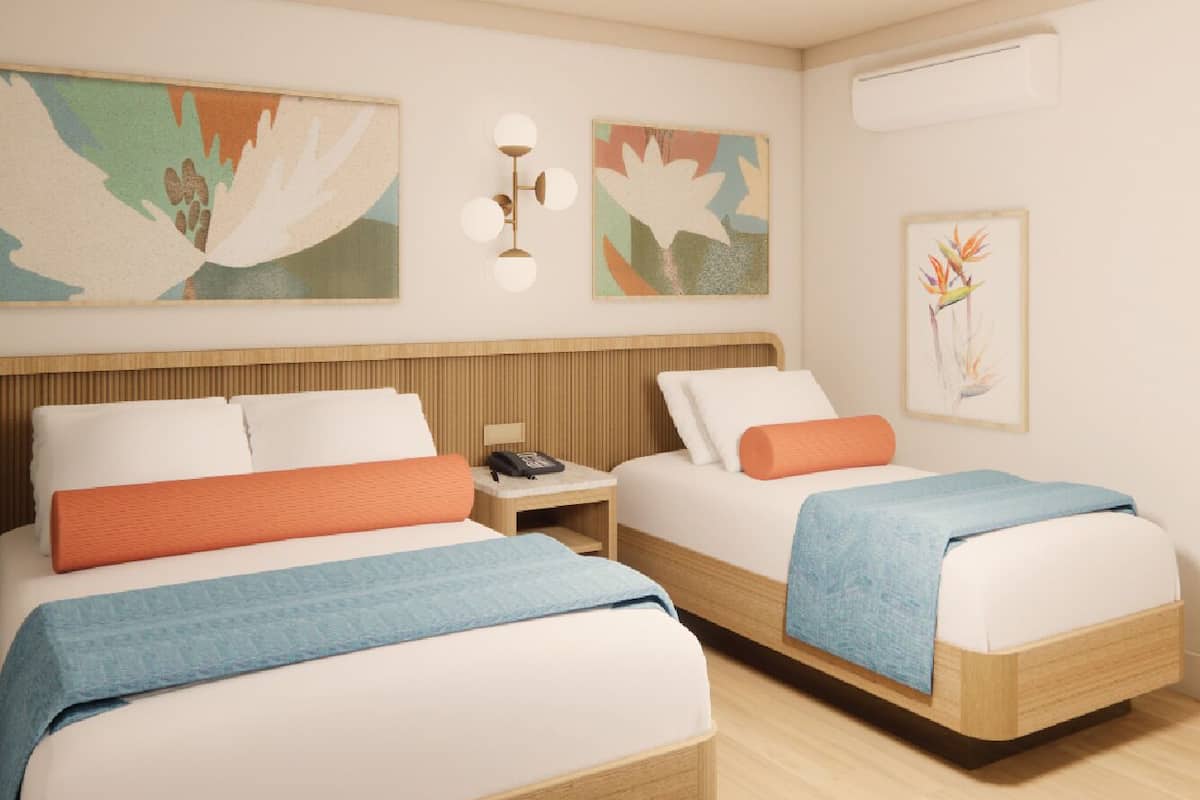 A hotel room with two beds, each with a wooden headboard, white bedding, and orange and blue throw pillows. There are wall-mounted light fixtures and framed artwork above the beds.