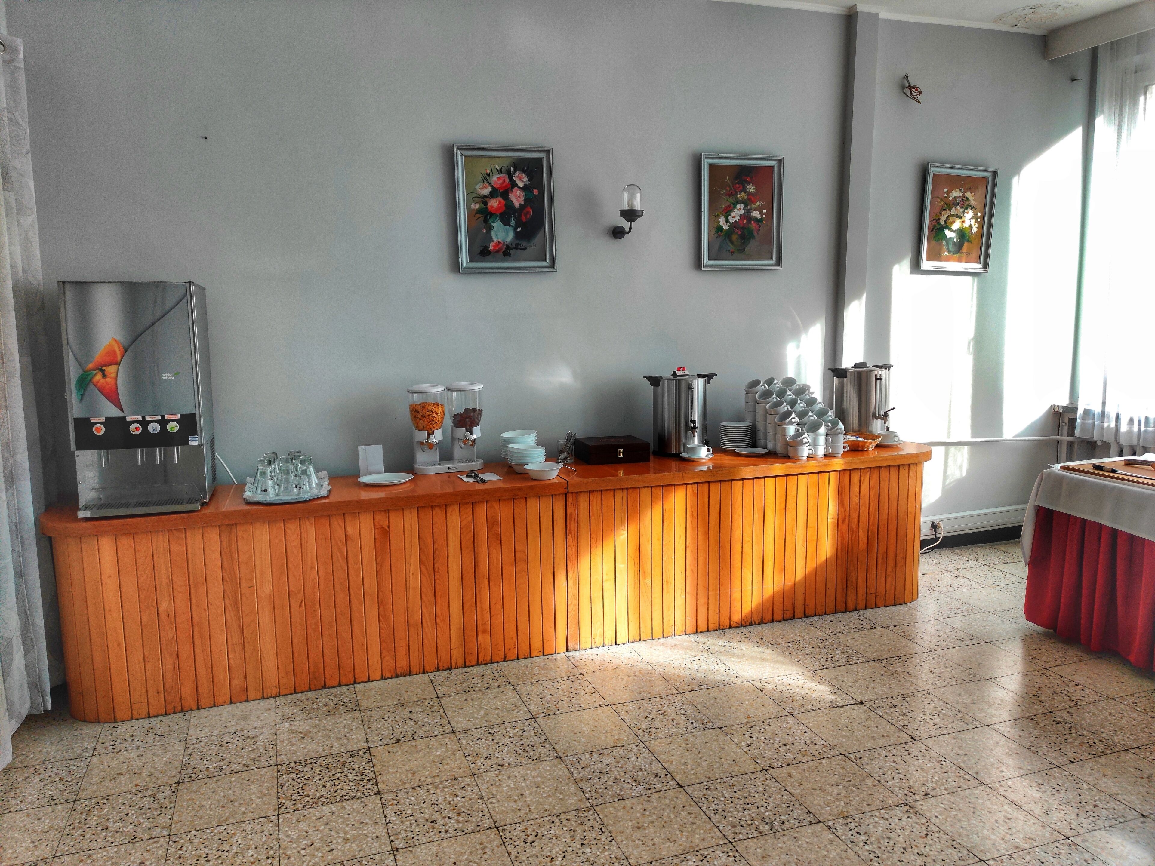 Een receptiegedeelte met een houten balie, een koffieapparaat en een waterdispenser. Er hangen bloemstilheden in lijsten aan de muur en een raam met gordijnen.