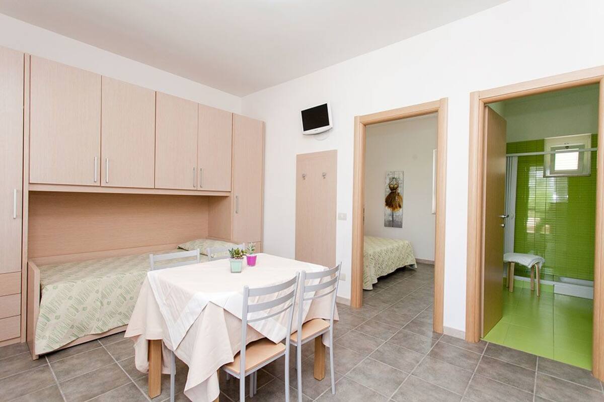 A compact room with a bed, a small table with chairs, a TV mounted on the wall, and a green door leading to another room.
