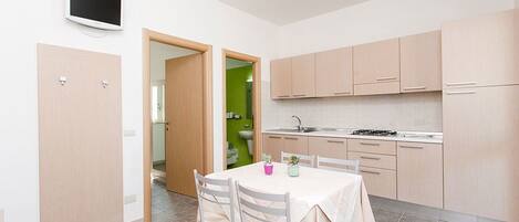A compact kitchen with light wood cabinets, a dining table with chairs, and a small TV mounted on the wall.